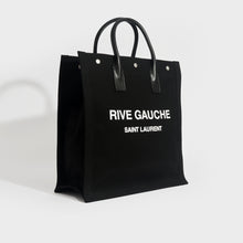 Load image into Gallery viewer, Side view of Saint Laurent Rive Gauche tote in black and canvas and white printed logo with black leather handles.