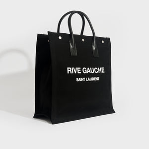 Side view of Saint Laurent Rive Gauche tote in black and canvas and white printed logo with black leather handles.